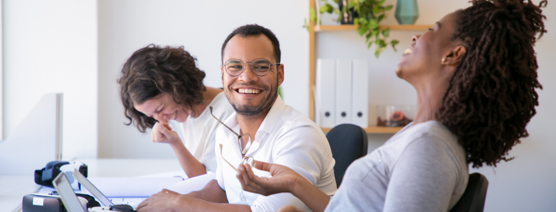 Three colleagues sitting together at a desk in a bright office, laughing heartily. A woman on the left looks down while giggling, a man in the centre smiles broadly while holding a pair of glasses, and a woman on the right throws her head back in laughter.